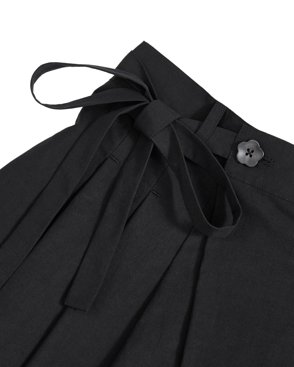 Close-up of the ASYMMETRIC CHIMA CULOTTE by Sundae School, black wrap pants with an asymmetric design, featuring a fabric waist tie in a bow, a flower-shaped black button on the waistband, and pleated detailing below the waistband.