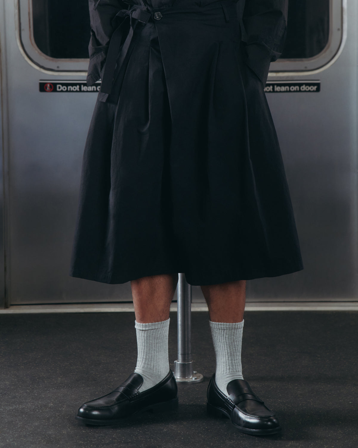 A person wearing the ASYMMETRIC CHIMA CULOTTE by Sundae School, paired with light gray socks and black loafers, stands inside a subway train with metal doors and a “Do not lean on door” sign in the background.