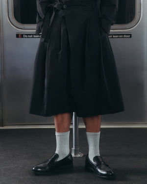 A person wearing the BLACK PLEATED HANBOK CULOTTES by Sundae School, paired with gray socks and black loafers, stands in a subway car with a metal pole and door in the background.