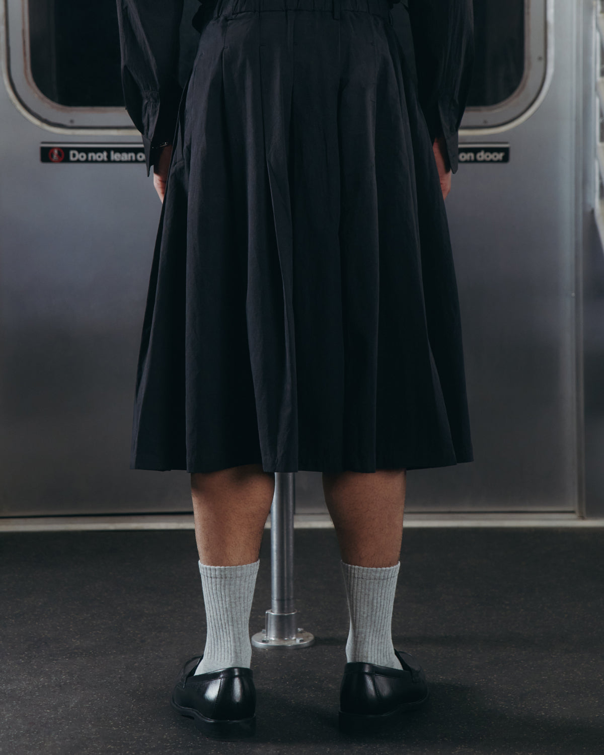 A person stands on a subway train, wearing the Sundae School ASYMMETRIC CHIMA CULOTTE—a flowing black skirt—paired with light gray socks and black shoes. Their back faces the camera, and a metal pole stands in front of them.