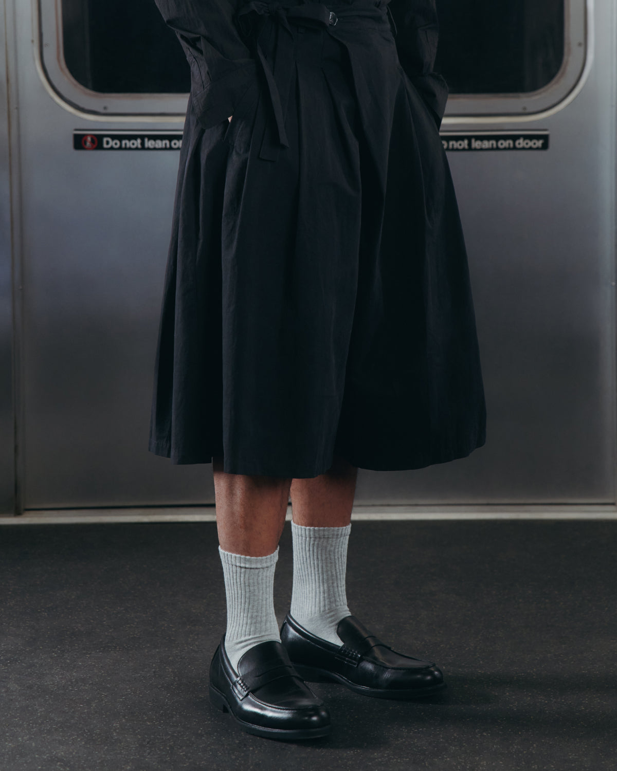 A person wearing the Sundae School ASYMMETRIC CHIMA CULOTTE, paired with white socks and black loafers, stands inside a subway train. Only their lower body is visible. A sign on the train door reads “Do not lean on door.”.