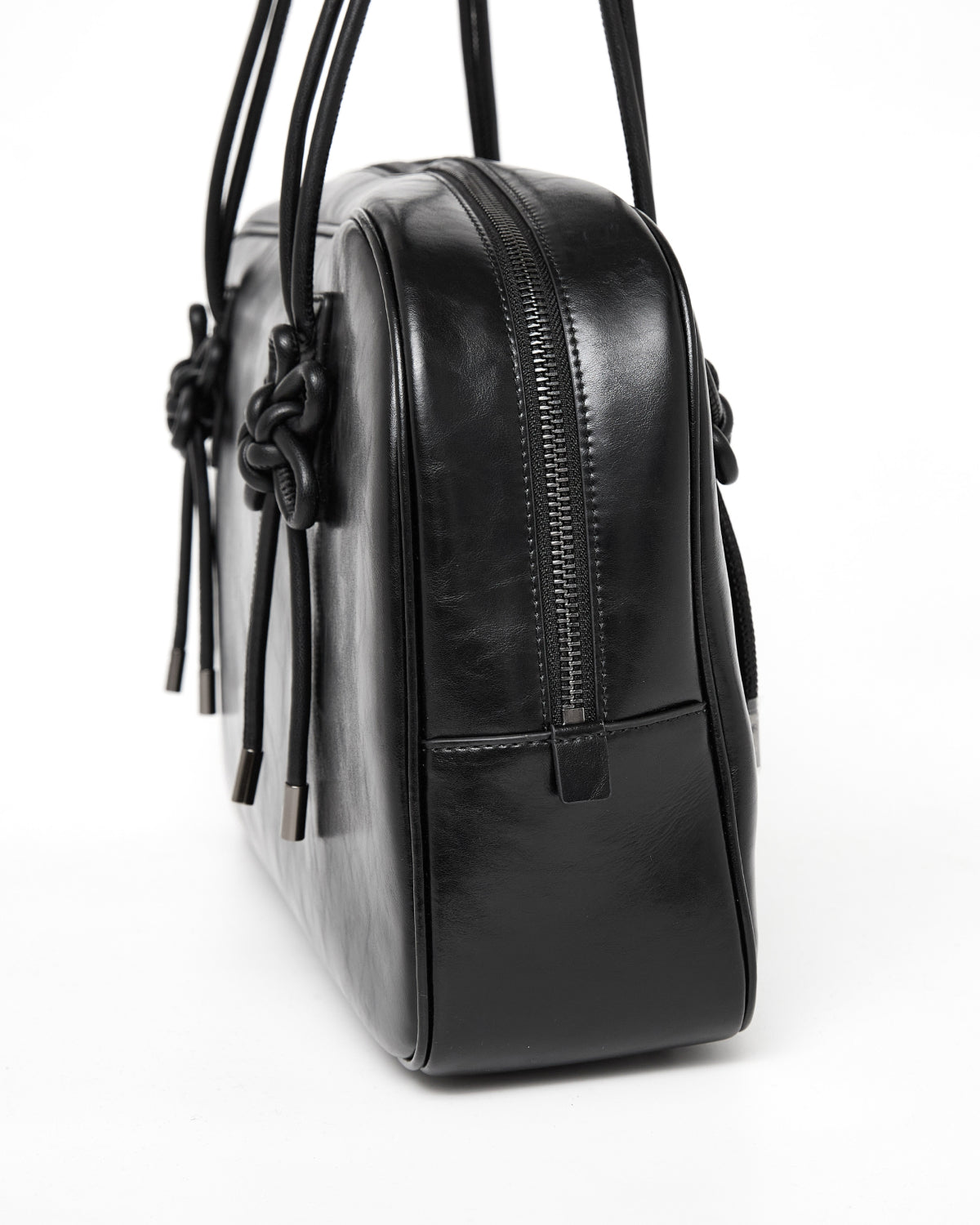 A close-up of the BLACK AUDREY BOWLING BAG by Sundae School, featuring a black leather exterior with a zipper closure, knotted leather straps, and metallic accents, photographed against a white background.