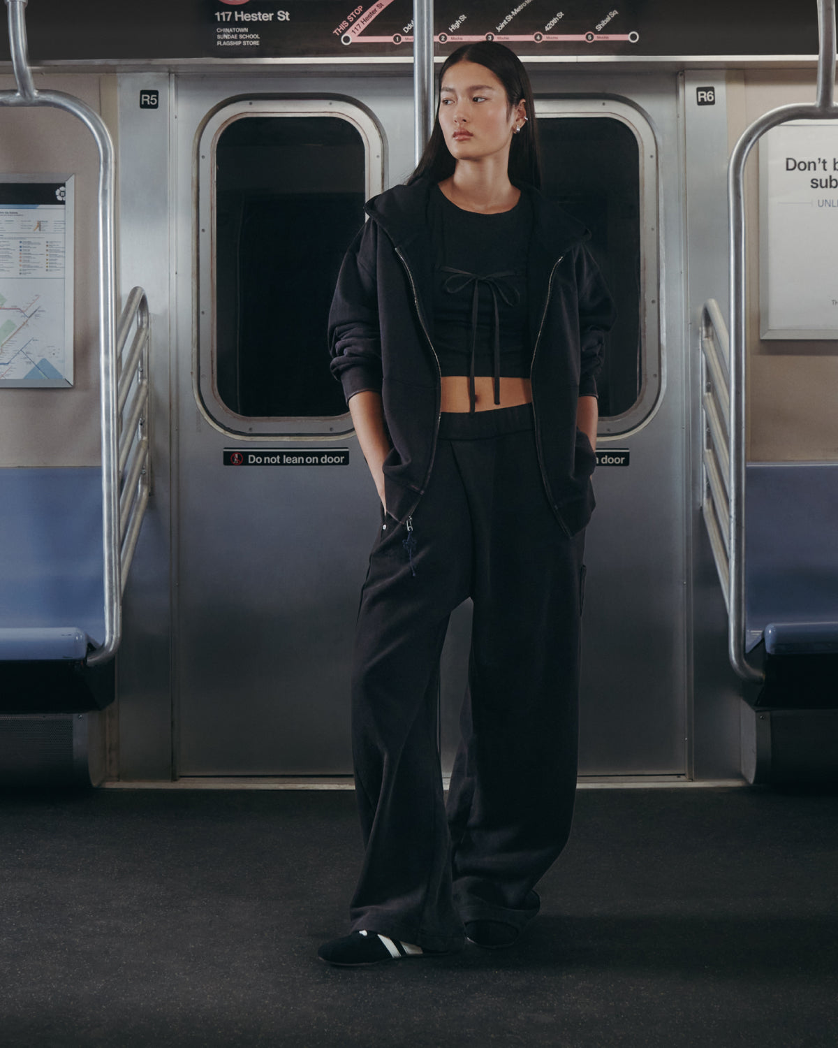 A person stands in a subway car wearing Sundae School’s BLACK WASH WRAP SWEAT BAJI. Their hands are in their pockets, and the subway seats around them are empty.