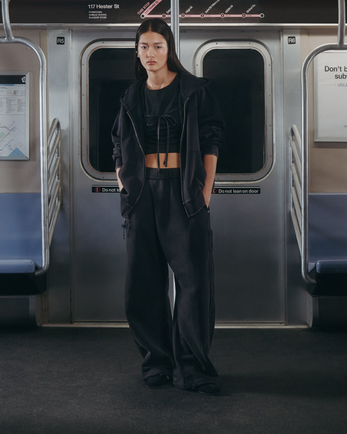 A person dressed in all-black casual attire, featuring Sundae School's BLACK WASH FLOWER KNOT ZIP UP with Hangul embroidery and a Maehwa knot zipper pull, stands in the center of an empty subway car, facing the camera with hands in their pockets. The station sign for 116th Street is visible above the doors.