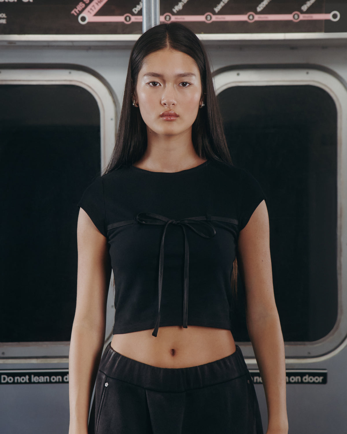 A woman with long dark hair wears the Sundae School BLACK TIE GORUM BABY TEE and black pants, standing by a subway window beneath 