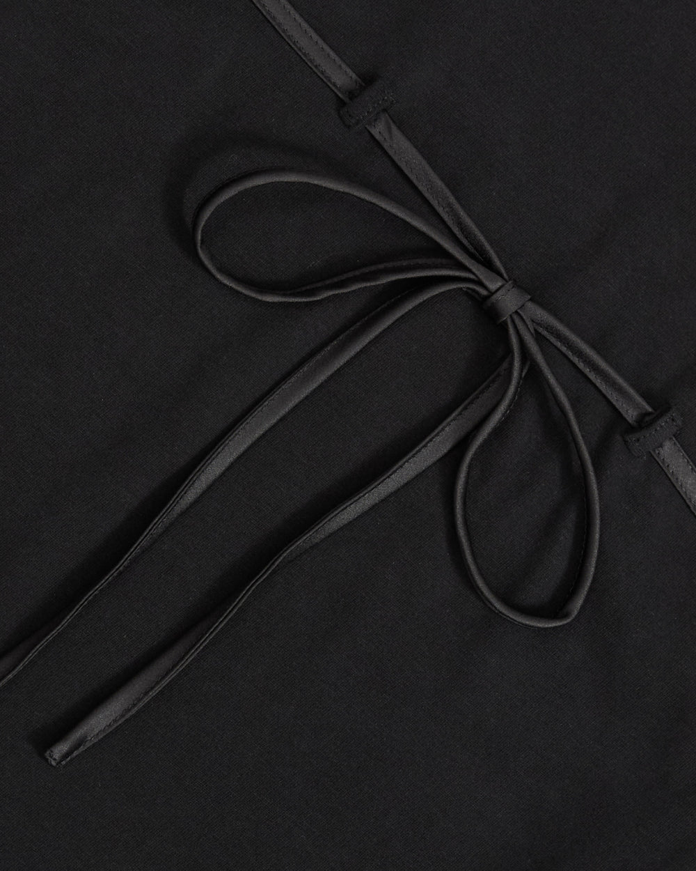 A close-up of the Sundae School BLACK TIE GORUM BABY TEE showcases a neatly tied, thin black ribbon bow set against a smooth, matte black fabric background. The elegant bow highlights the modern, minimalist design of this form-fitting women’s shirt.