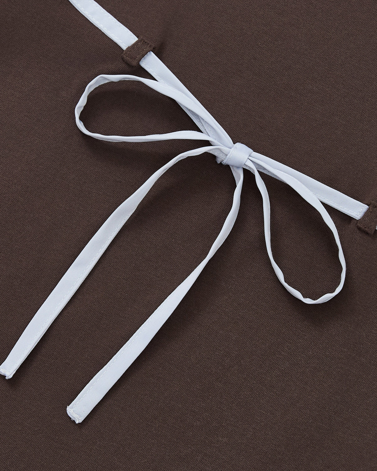 A white ribbon is tied in a chest-strap bow on the BROWN TIE GORUM BABY TEE by Sundae School, displayed against dark brown fabric.
