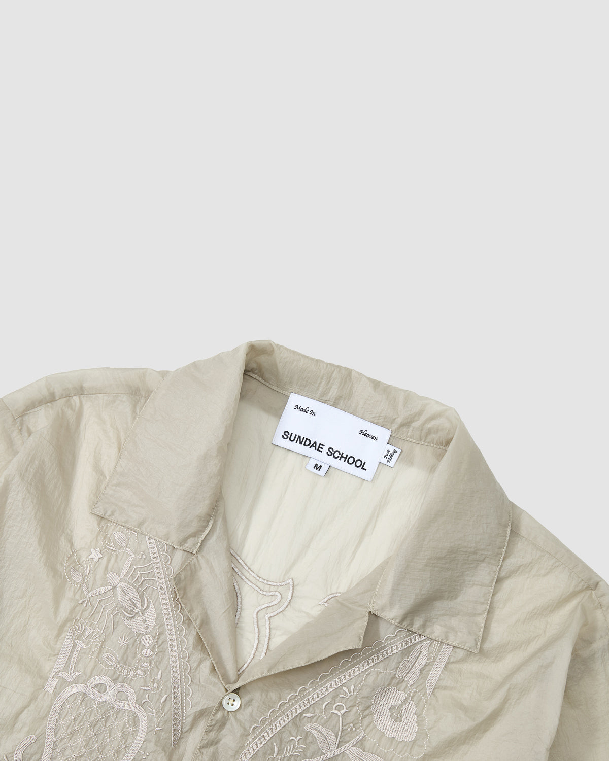 The BARONG SHIRT by Sundae School is a beige collared shirt with detailed embroidery near the buttons and an inside label marked size M. Inspired by the Filipino Barong Tagalog, it’s displayed partially folded on a light gray background.
