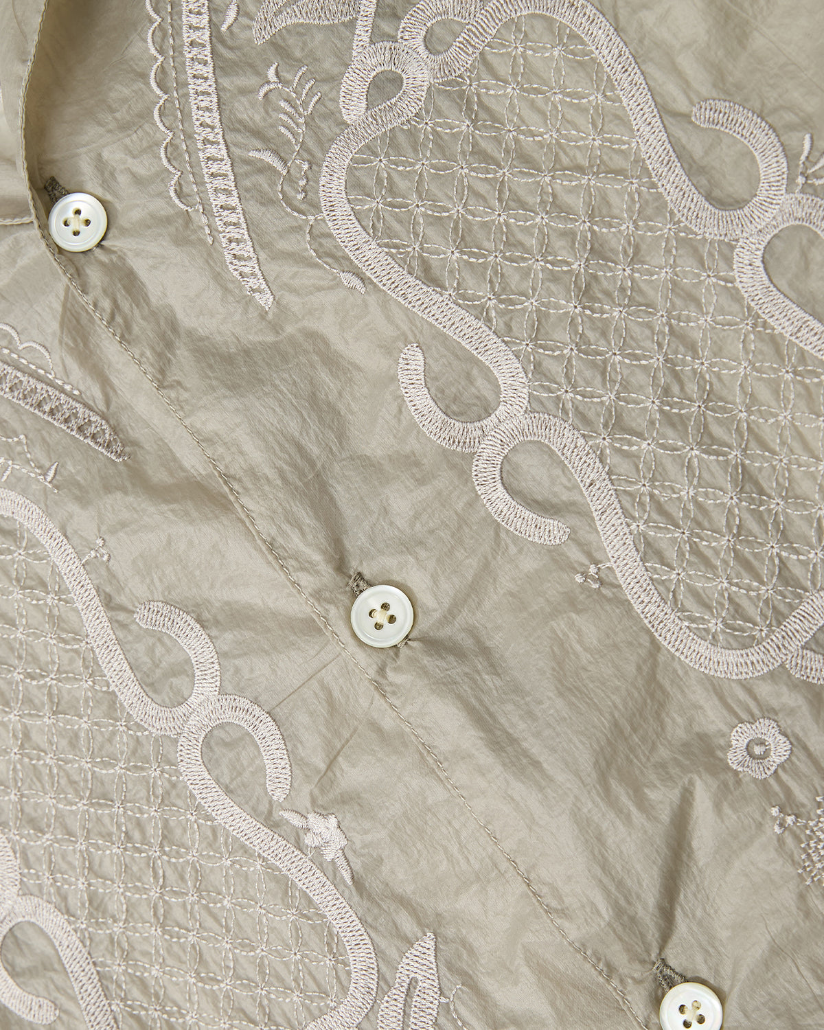 Close-up of a beige Sundae School BARONG SHIRT fabric, featuring intricate white embroidery with geometric grids, swirls, small floral motifs, and three white buttons in a classic Filipino silhouette.