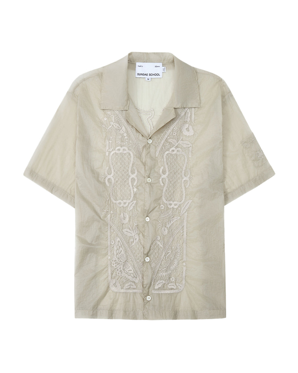 The BARONG SHIRT by Sundae School is a light beige short-sleeve button-up featuring intricate embroidery on the front, a camp collar, and design details inspired by the classic Filipino Barong Tagalog silhouette. This relaxed-fit shirt includes a visible Sundae School label inside the collar and subtle references to traditional craftsmanship.