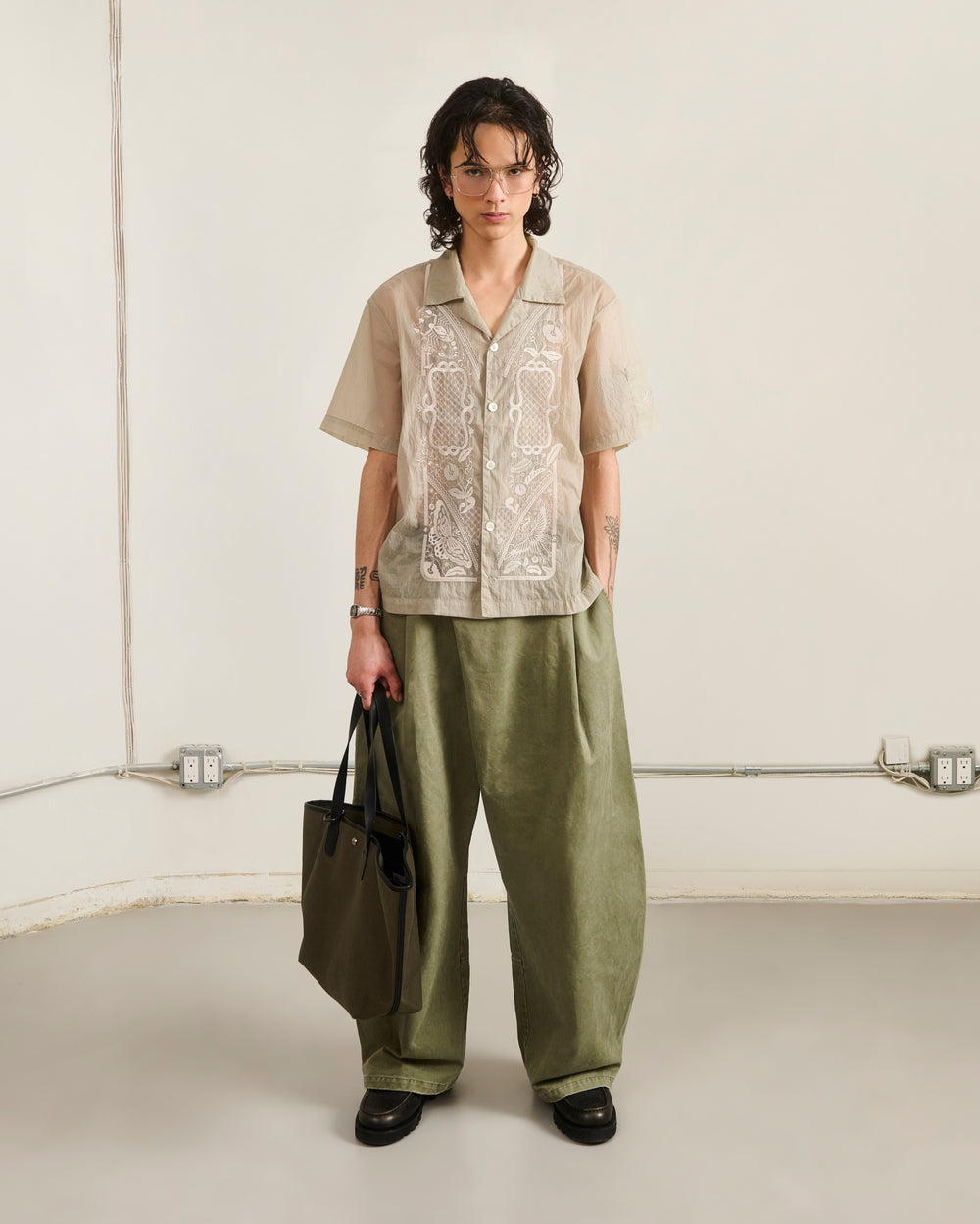 A person with short, wavy hair stands indoors wearing glasses, loose olive green pants, black shoes, and holding a green tote bag. They sport Sundae School's BARONG SHIRT—nylon, translucent, and embroidered. The background is minimal.