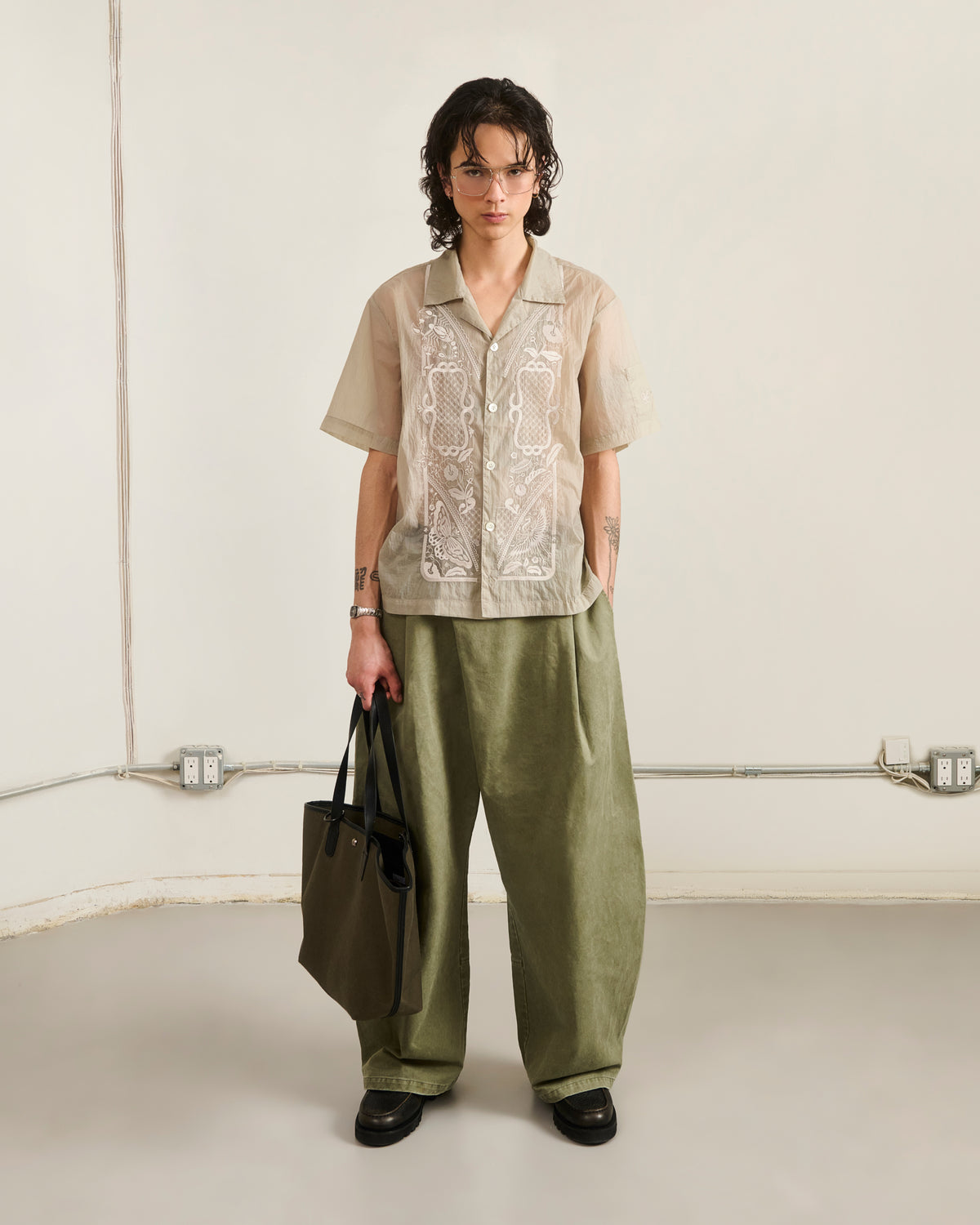 A person with short, wavy hair stands indoors wearing glasses, loose olive green pants, black shoes, and holding a green tote bag. They sport Sundae School's BARONG SHIRT—nylon, translucent, and embroidered. The background is minimal.