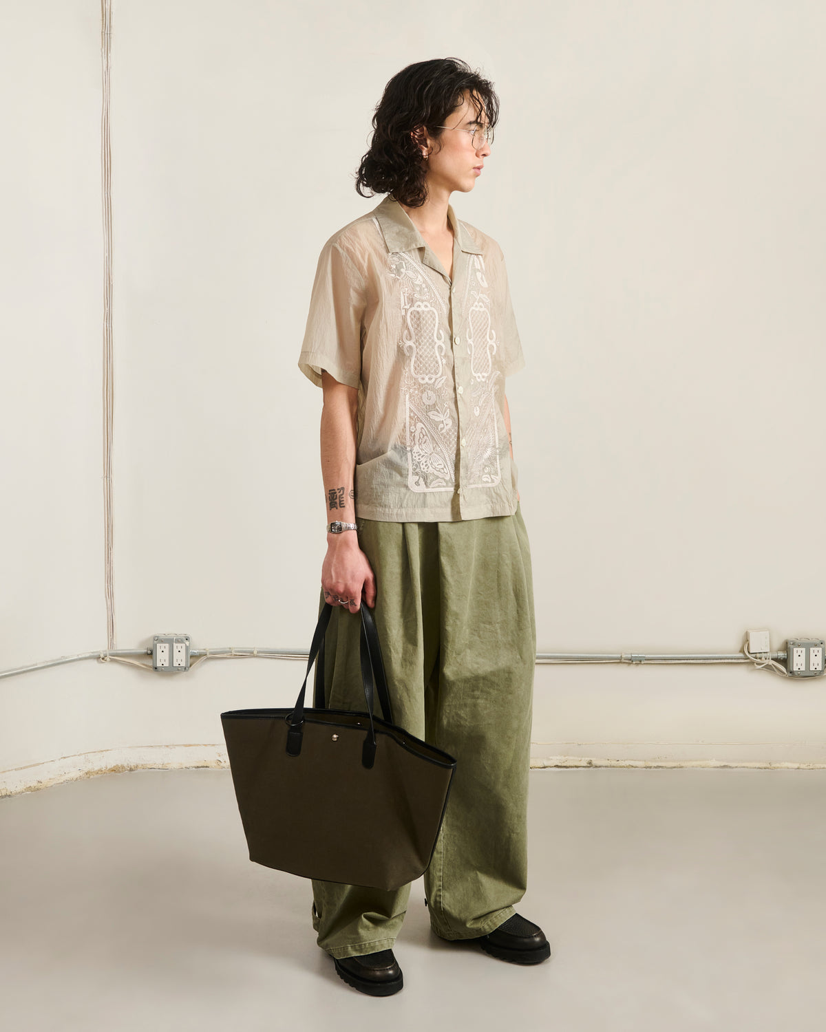 A person with wavy hair stands indoors wearing the Sundae School BARONG SHIRT, sheer beige and inspired by the Barong Tagalog, paired with loose olive green pants, black shoes, and holding a large olive tote bag. Plain background with visible outlets.
