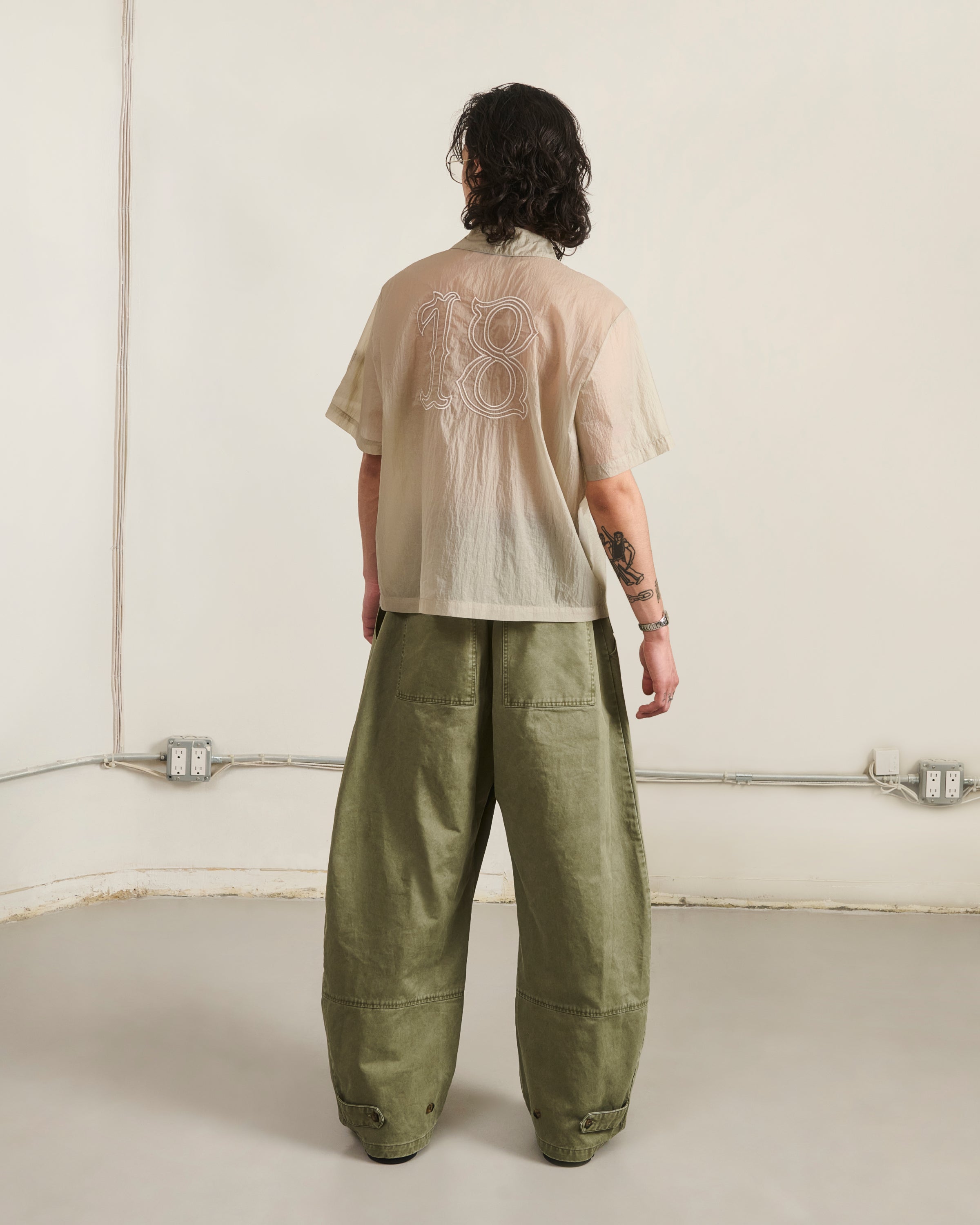 In a minimalist room, a person with shoulder-length wavy hair and left arm tattoos wears Sundae School's BARONG SHIRT—nylon, translucent, 