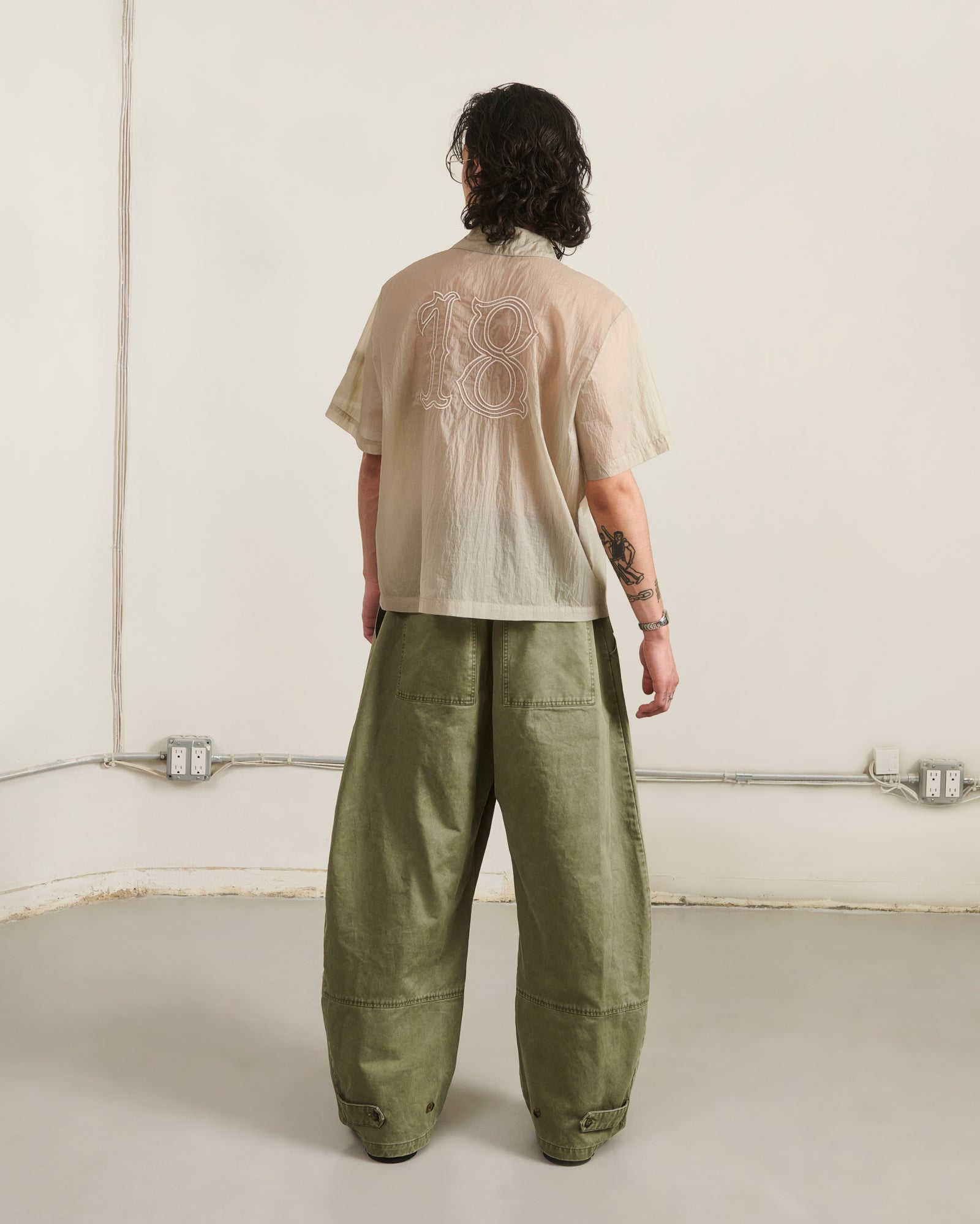 In a minimalist room, a person with shoulder-length wavy hair and left arm tattoos wears Sundae School's BARONG SHIRT—nylon, translucent, "18" on the back—paired with loose olive pants, creating a striking Filipino silhouette.