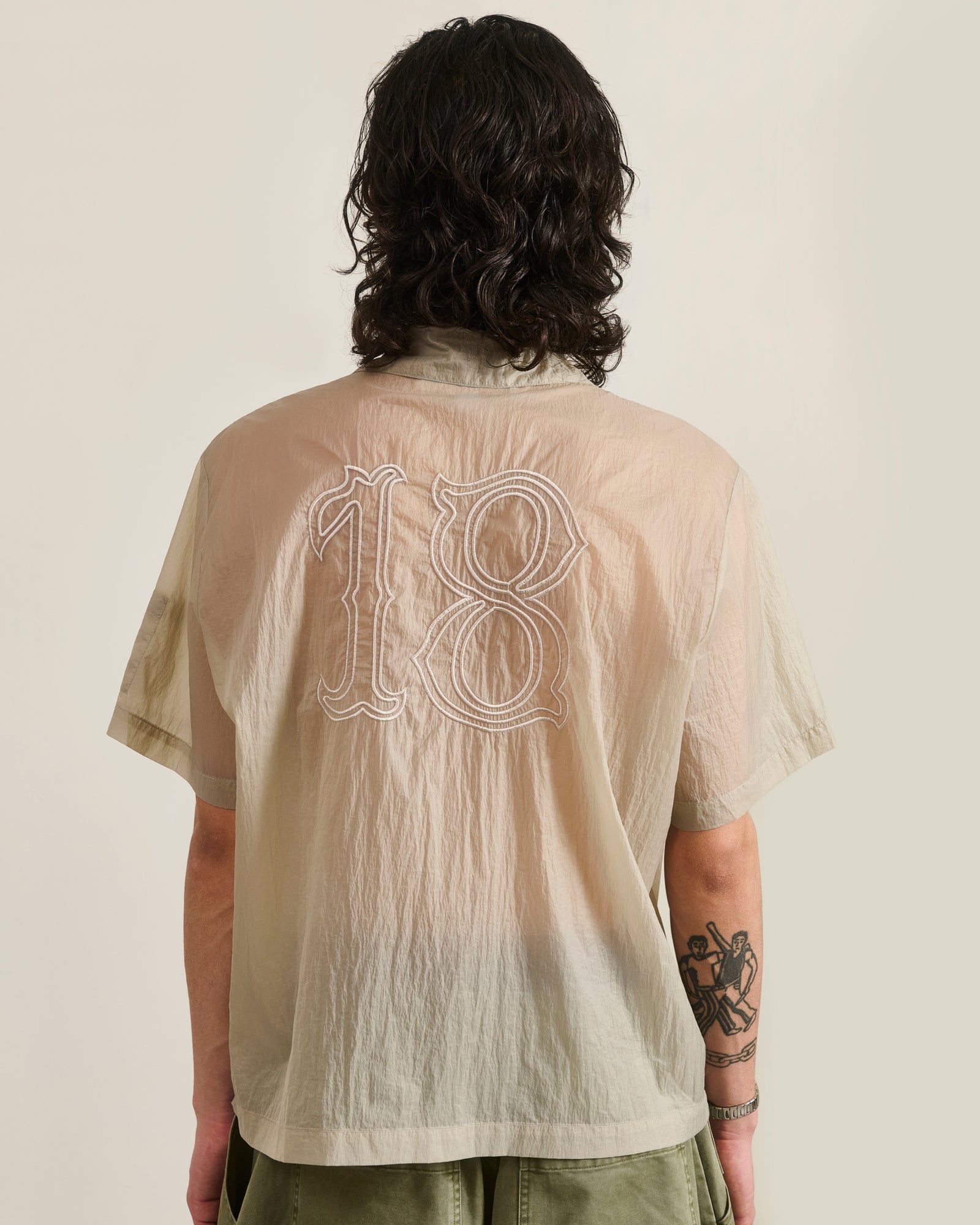 A person with long dark hair, wearing the Sundae School BARONG SHIRT—a sheer beige nylon shirt in a Filipino silhouette with an embroidered number 18 on the back—stands facing away, revealing a left forearm tattoo.