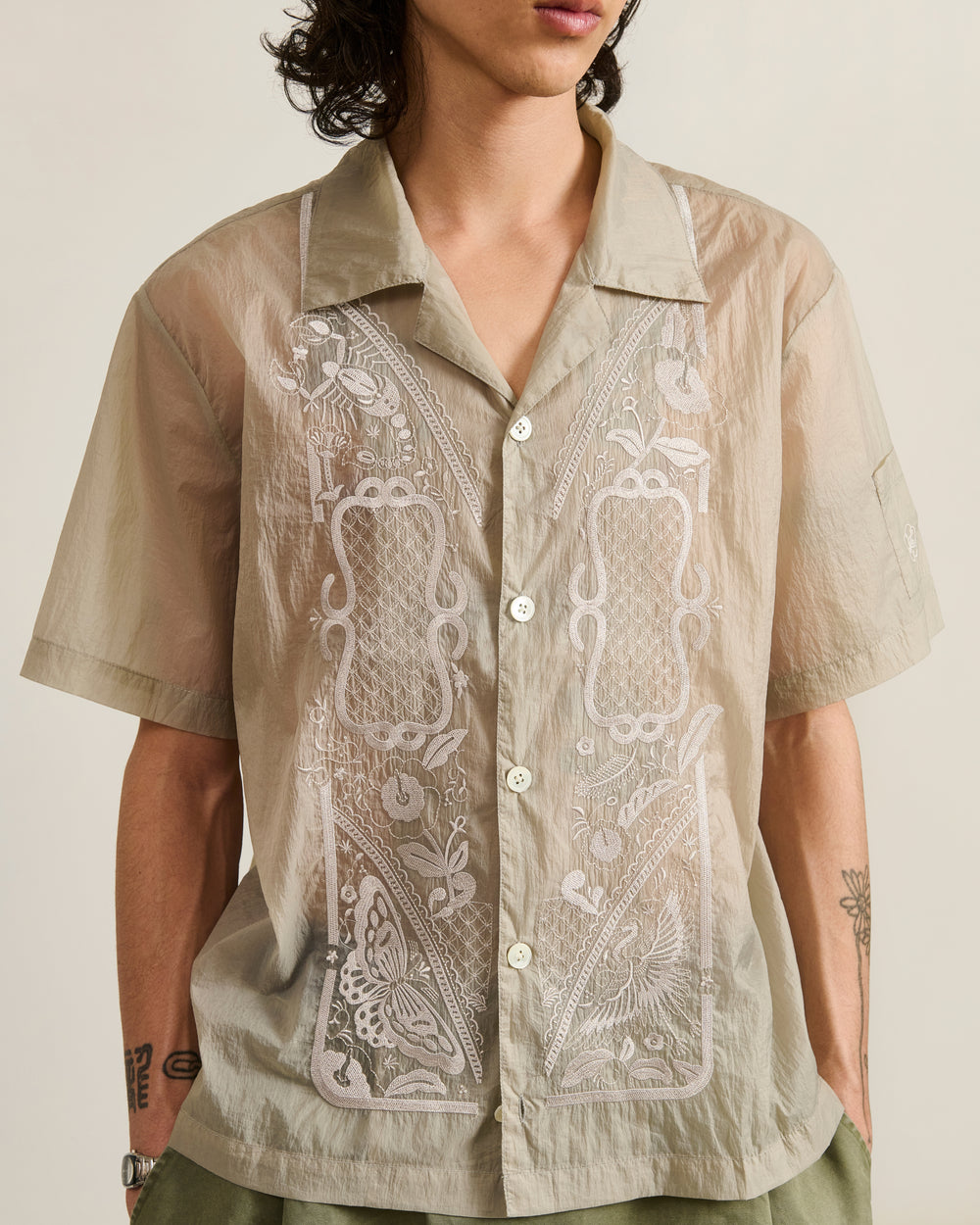 A person with wavy, shoulder-length hair is wearing the Sundae School BARONG SHIRT, a sheer light beige short-sleeve button-up featuring intricate white floral and geometric embroidery. Their face is partially obscured.