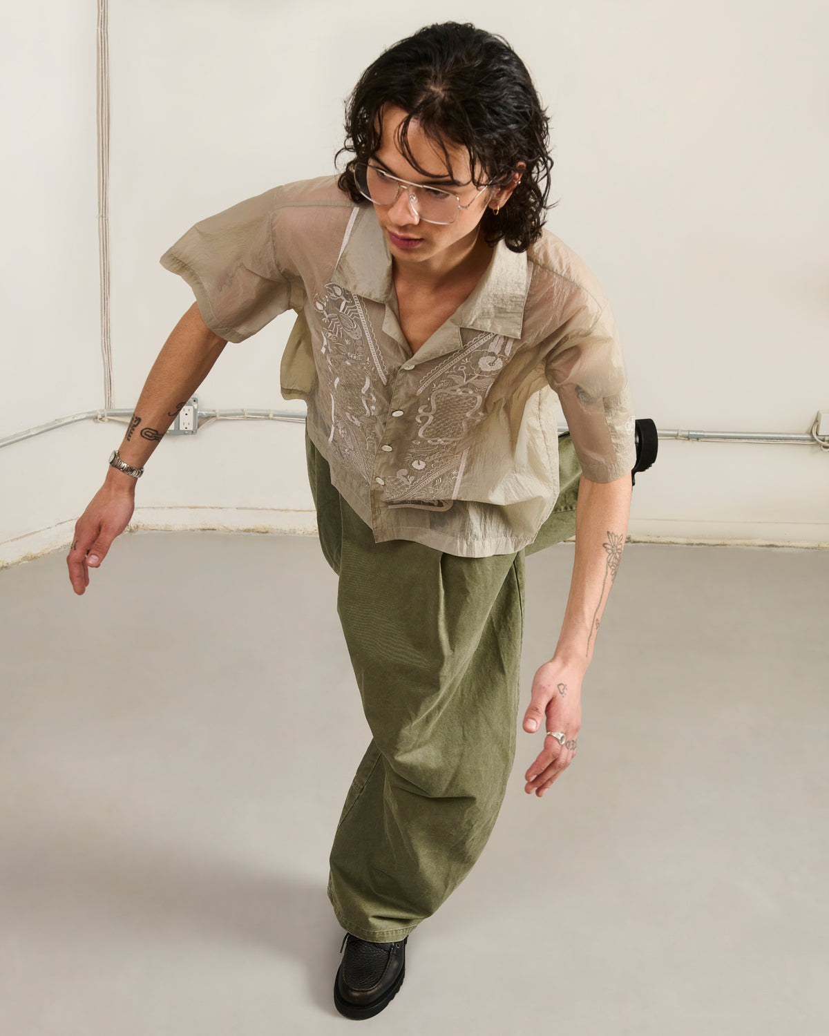 A person with wavy hair and glasses wears the Sundae School BARONG SHIRT—nylon, translucent, Barong Tagalog–inspired—paired with loose green pants, leaning forward on a light gray floor against a plain wall with visible wiring.