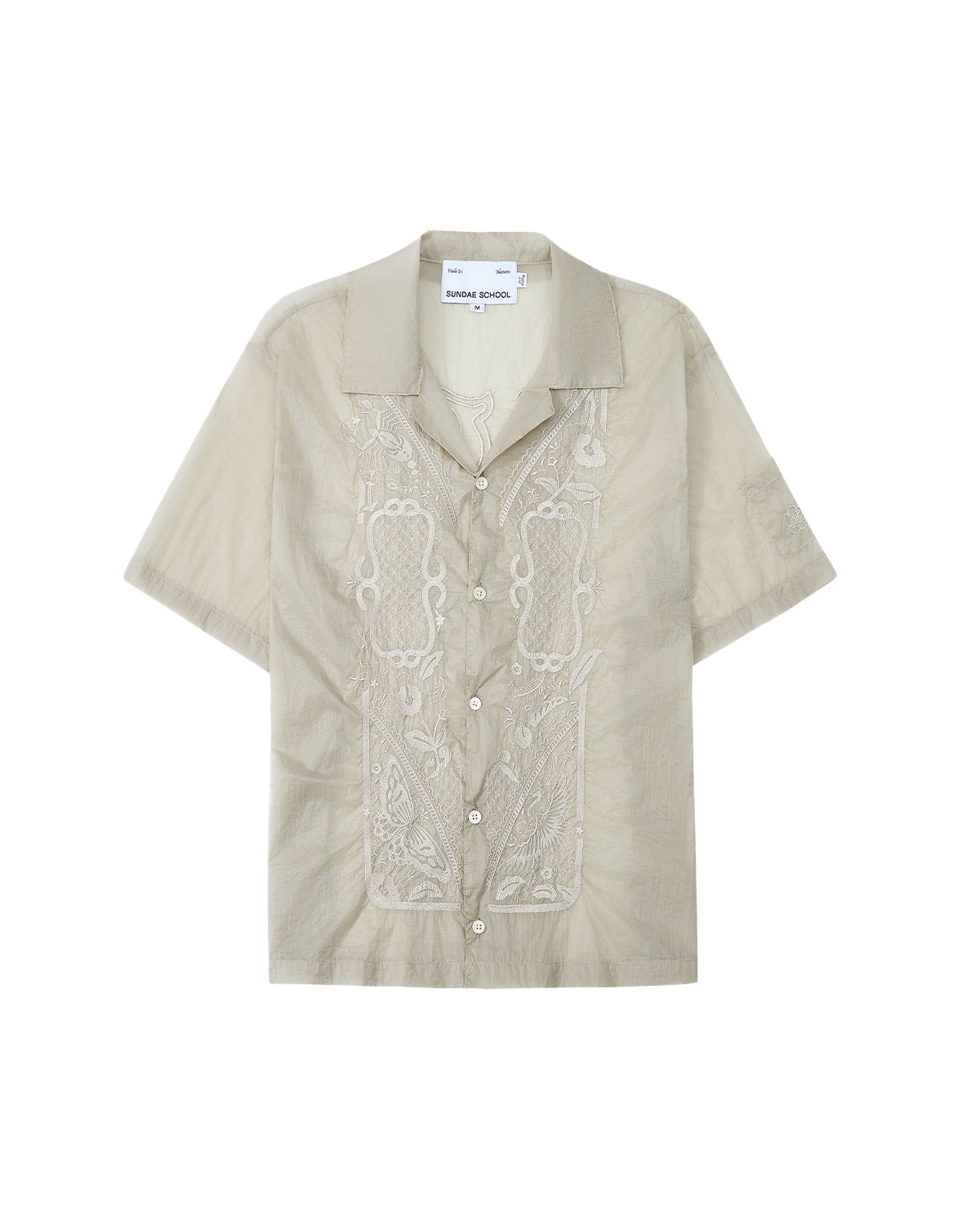 The BARONG SHIRT by Sundae School is a light beige short-sleeve button-up featuring intricate embroidery on the front, a camp collar, and design details inspired by the classic Filipino Barong Tagalog silhouette. This relaxed-fit shirt includes a visible Sundae School label inside the collar and subtle references to traditional craftsmanship.