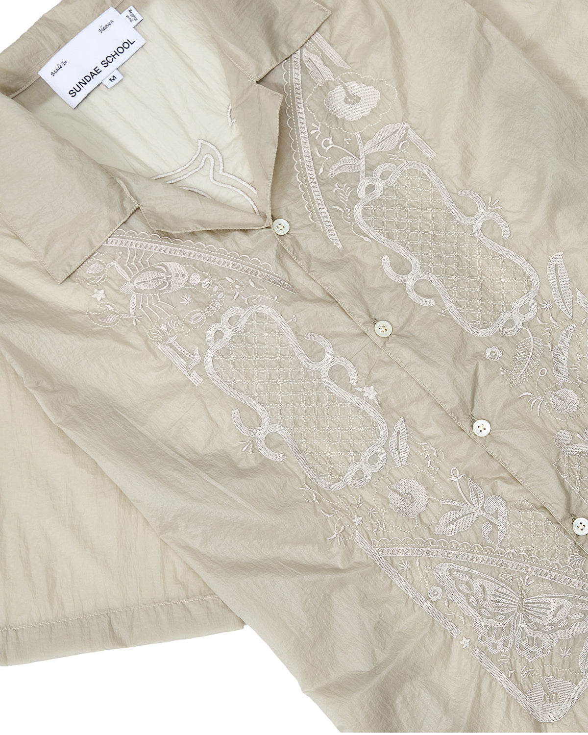 A beige BARONG SHIRT by Sundae School, inspired by the Filipino silhouette and crafted from translucent nylon with a subtle sheen reminiscent of the traditional Barong Tagalog. This short-sleeve shirt features delicate floral embroidery, front buttons, and includes an interior SUNDAY SCHOOL label with a size M tag.