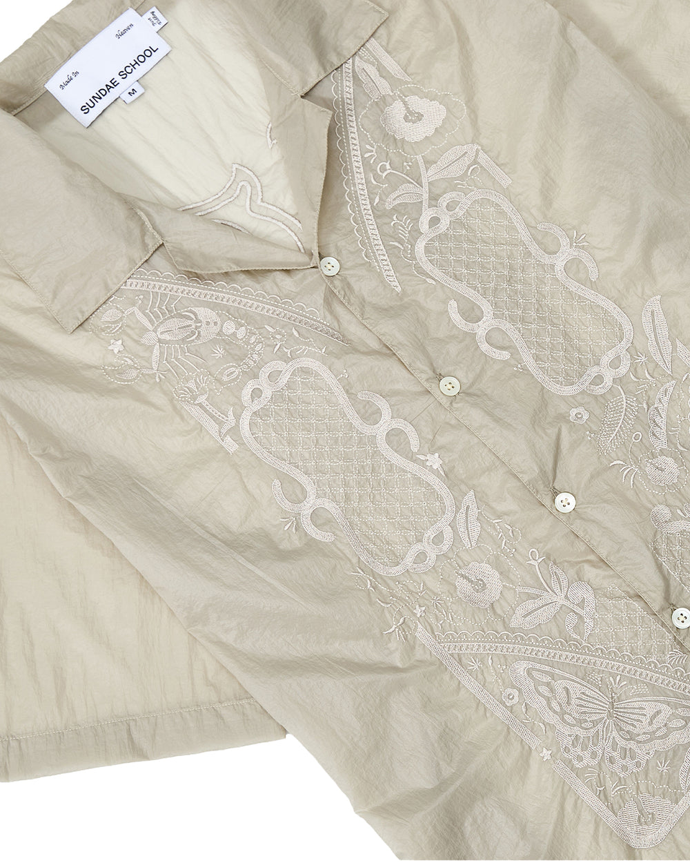 A beige BARONG SHIRT by Sundae School, inspired by the Filipino silhouette and crafted from translucent nylon with a subtle sheen reminiscent of the traditional Barong Tagalog. This short-sleeve shirt features delicate floral embroidery, front buttons, and includes an interior SUNDAY SCHOOL label with a size M tag.