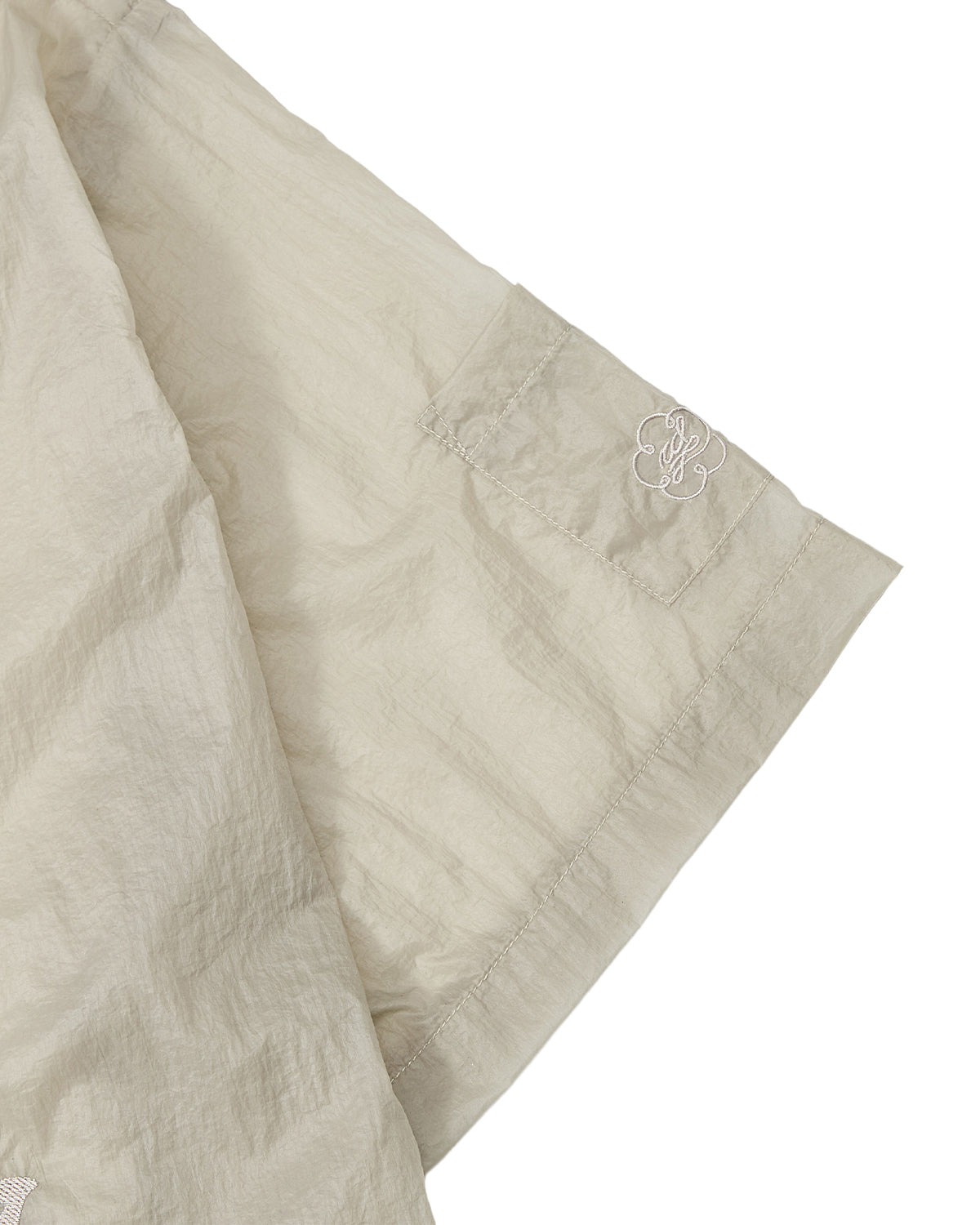A close-up of a light beige sleeve from the BARONG SHIRT by Sundae School, featuring a subtle white embroidered logo near the hem. The lightweight, slightly crinkled fabric evokes the translucent nylon material often used in modern reinterpretations of the Barong Tagalog.