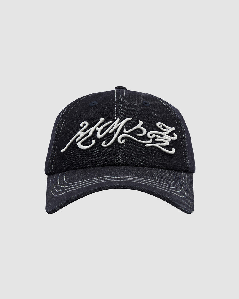 The INDIGO DAENGGI CAP by Sundae School is a black denim 6-panel cap with adjustable fit, white stylized script embroidery on the front, and crisp visible white stitching along the brim and panels.