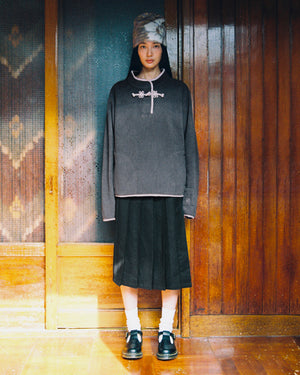 In front of wooden doors indoors, a person looks directly at the camera, wearing Sundae School’s CHARCOAL BUTTERFLY FLEECE PULLOVER—a gray mock-neck polar fleece with tteokbokki toggles—paired with a gray hat, black pleated skirt, white socks, and black shoes.