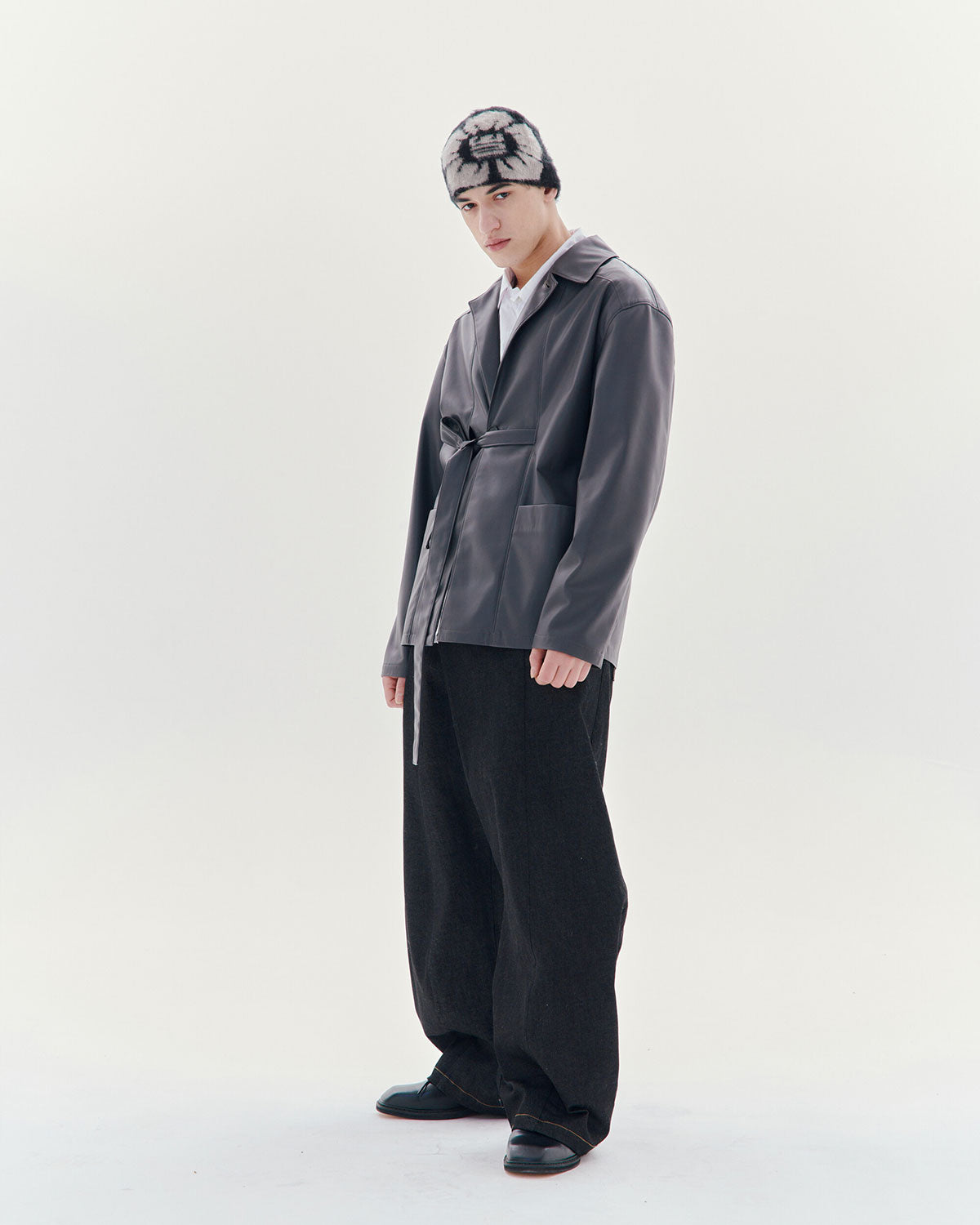 A person stands against a white background wearing a gray jacket, wide black pants, black shoes, and the Sundae School Black Ddul Beanie, featuring a black-and-white pattern, looking slightly down with a relaxed posture.