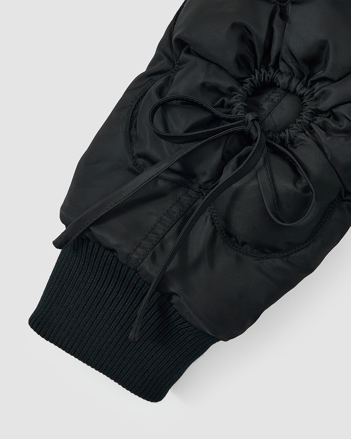 Close-up of the BLACK FLOWER CHANNELED BOMBER by Sundae School, showing a black puffy sleeve with ruched detailing, a tied bow at the wrist, and a ribbed knit cuff against a plain light gray background.