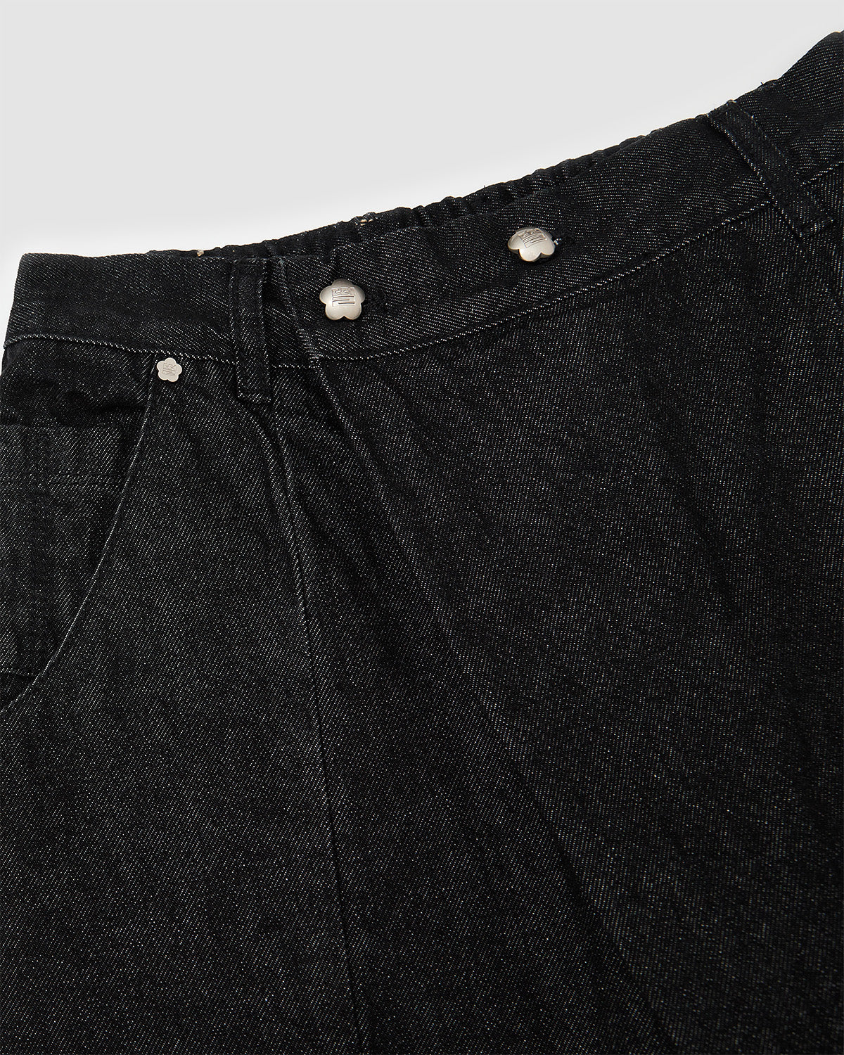 Close-up detail of black hanbok denim waistband showing silver flower-shaped snap buttons and asymmetric front closure design, highlighting custom hardware and button placement