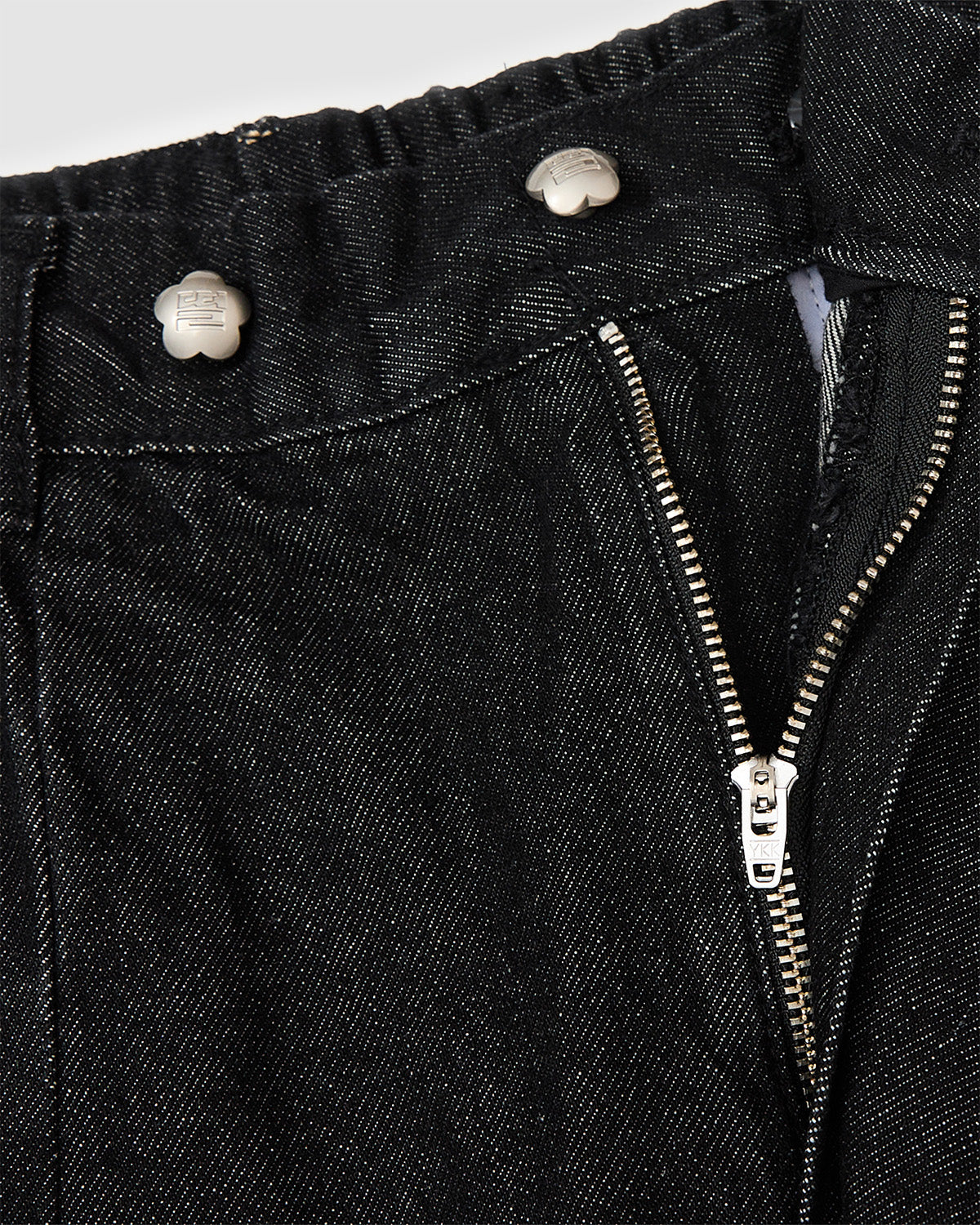 Close-up detail of black hanbok denim showing partially open zipper closure and silver flower-shaped snap button hardware, highlighting construction details and custom closure system