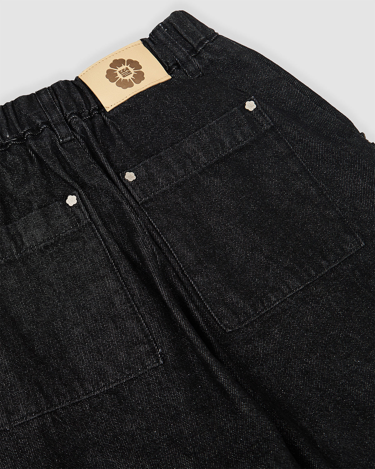Close-up detail of Sundae School tan leather brand patch with embossed flower logo on black hanbok denim waistband, showing back pocket construction with silver snap button hardware