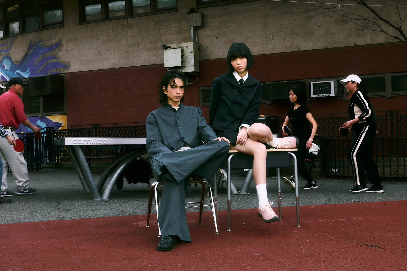 A fashion editorial photograph featuring models in SUNDAE SCHOOL SAILOR COLLAR UNIFORM, APRON SKIRT and STAND COLLAR UNIFORM JACKET and PLEATED UNIFORM PANTS posing on metal chairs in Chinatown, NYC. The central models wear dark gray and black traditional-inspired jackets and trousers, with one model sitting on a table wearing a black blazer, white shirt, and black tie.