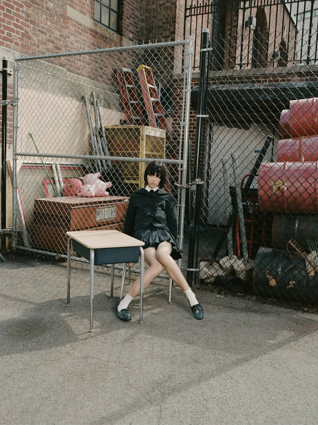 A person wearing SAILOR COLLAR UNIFORM JACKET and APRON SKIRT sits on a small desk in an urban alleyway with a chain-link fence, brick wall, and stacked red barrels in the background.