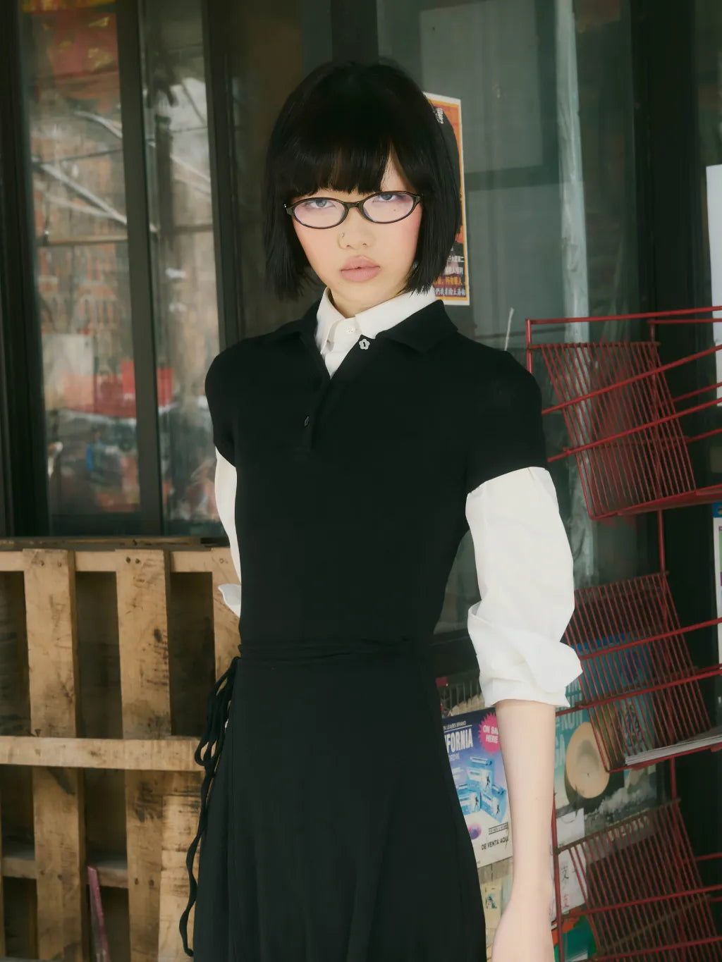 A model with short dark hair and glasses wearing CINDY POLO DRESS over a white long-sleeve shirt, standing in front of a shop window with wooden crates and a red wire rack in the background.