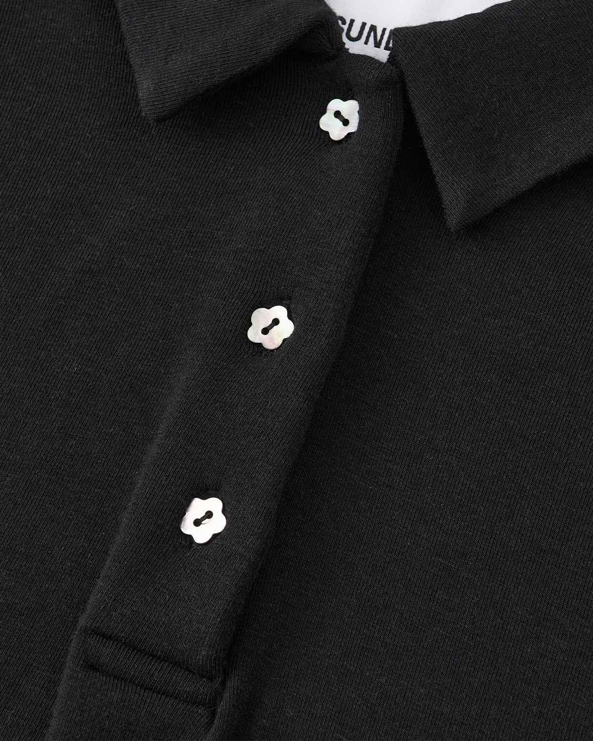 Close-up of the BLACK CINDY POLO DRESS by Sundae School, featuring a black collar and three iridescent, flower-shaped buttons on the placket—details that evoke classic sophistication. The edge of a white shirt label is partially visible inside the collar.