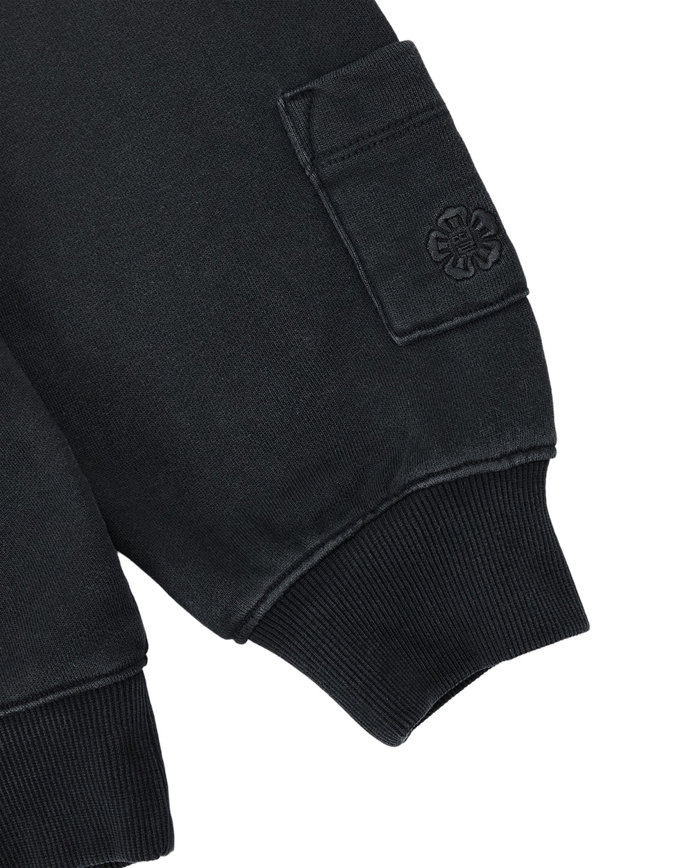A close-up of the black ribbed cuff on the sleeve of the FADE BLACK COCOON HOODIE by Sundae School, with a small pocket adorned with an embossed flower-like emblem.