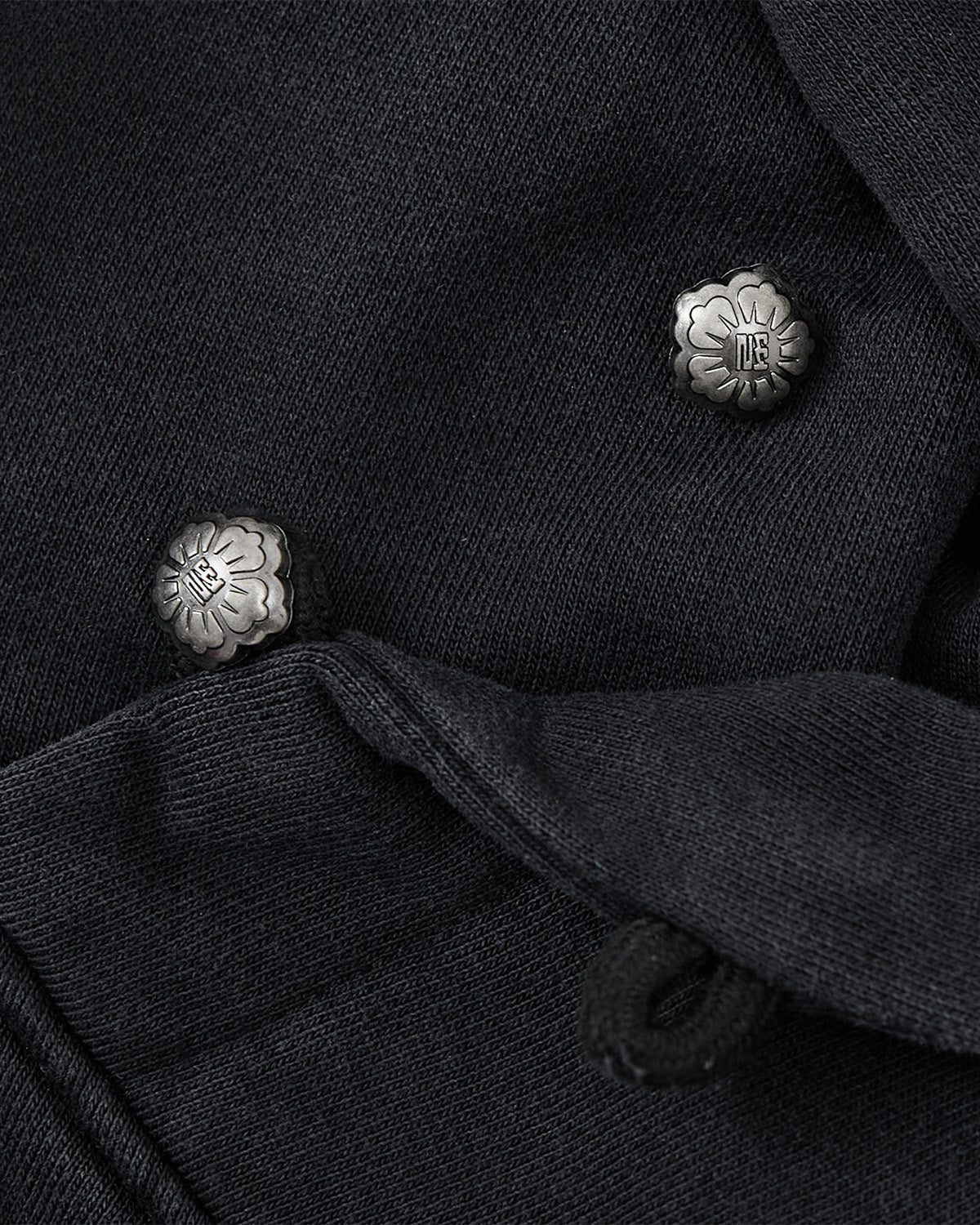 Close-up of two silver floral metal buttons on dark fabric, with one button partly covered by a folded section of the material—an elegant detail that perfectly complements Sundae School’s FADE BLACK COCOON HOODIE.
