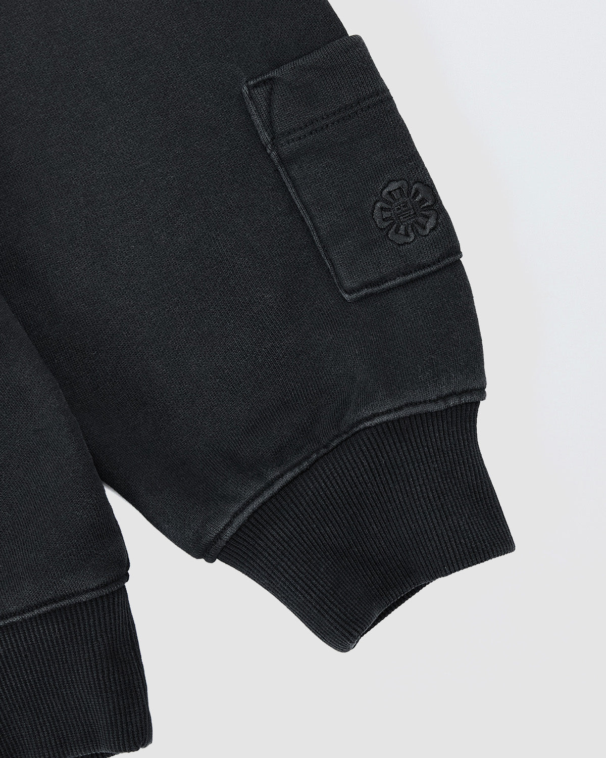 A close-up of the Sundae School FADE BLACK COCOON HOODIE shows a ribbed cuff, rectangular pocket with embroidered four-leaf clover, and cocoon-core appeal from the soft, textured fabric.