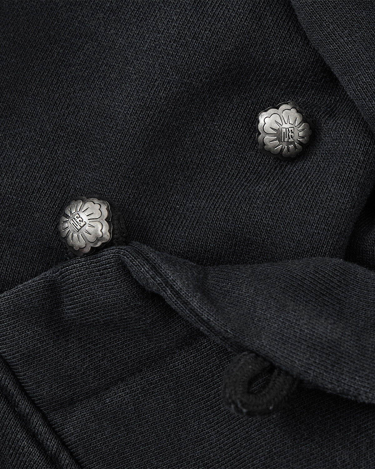 Close-up of the FADE BLACK COCOON HOODIE by Sundae School, showcasing black textured fabric and two ornate metallic floral buttons with engraved letters, evoking a cocoon-core vibe and a unique, limited-edition feel.