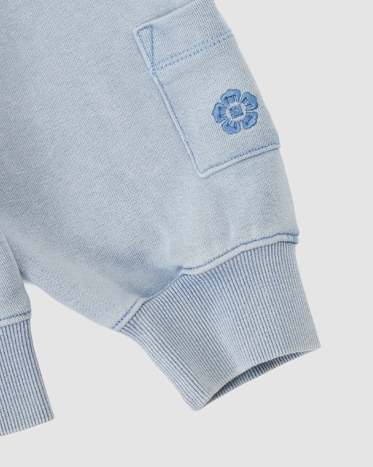 Close-up of Sundae School’s SKY BLUE COCOON HOODIE showing a light blue knit sleeve with ribbed cuff, peret sleeves, small pocket, and embroidered blue flower near the pocket for a soft cocoon-core vibe.