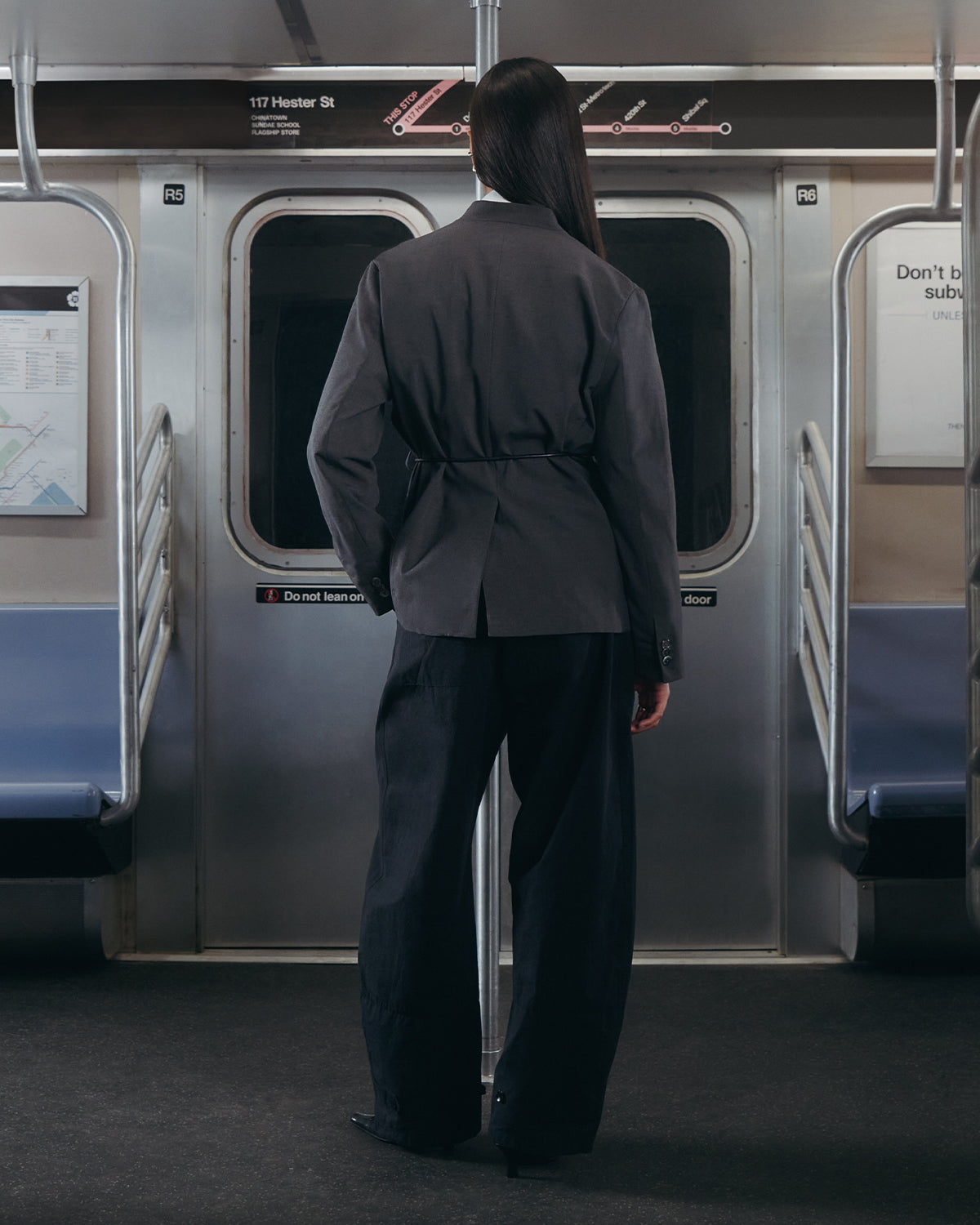 A person with long dark hair, wearing Sundae School's COLLARLESS TIE BLAZER and a vegan leather belt, stands facing the closed doors inside an empty subway train with maps on the walls.