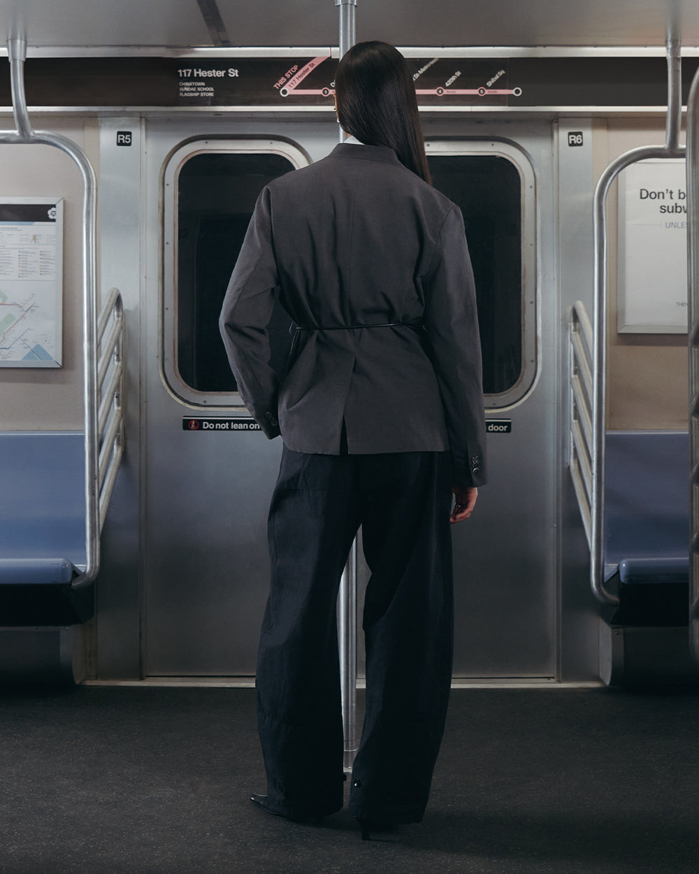 A person with long dark hair, dressed in Sundae School’s BLACK FLOWER NORIGAE LINEN TROUSERS, stands facing the subway doors inside a mostly empty subway car, with subway maps and signs visible on the walls.