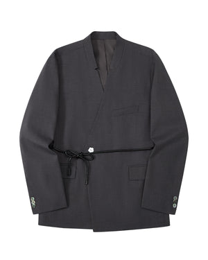 The COLLARLESS TIE BLAZER by Sundae School is a dark gray blazer with an unstructured drape and a shawl collar, designed without buttons. It features a single chest pocket, two front flap pockets, and comes with a thin vegan leather belt that ties at the waist.