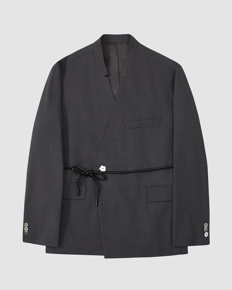 The Sundae School COLLARLESS TIE BLAZER is a dark gray, collarless summer wool blazer with a wrap front, chest pocket, two flap pockets, buttoned cuffs, and a vegan leather waist belt, shown on a plain light gray background.