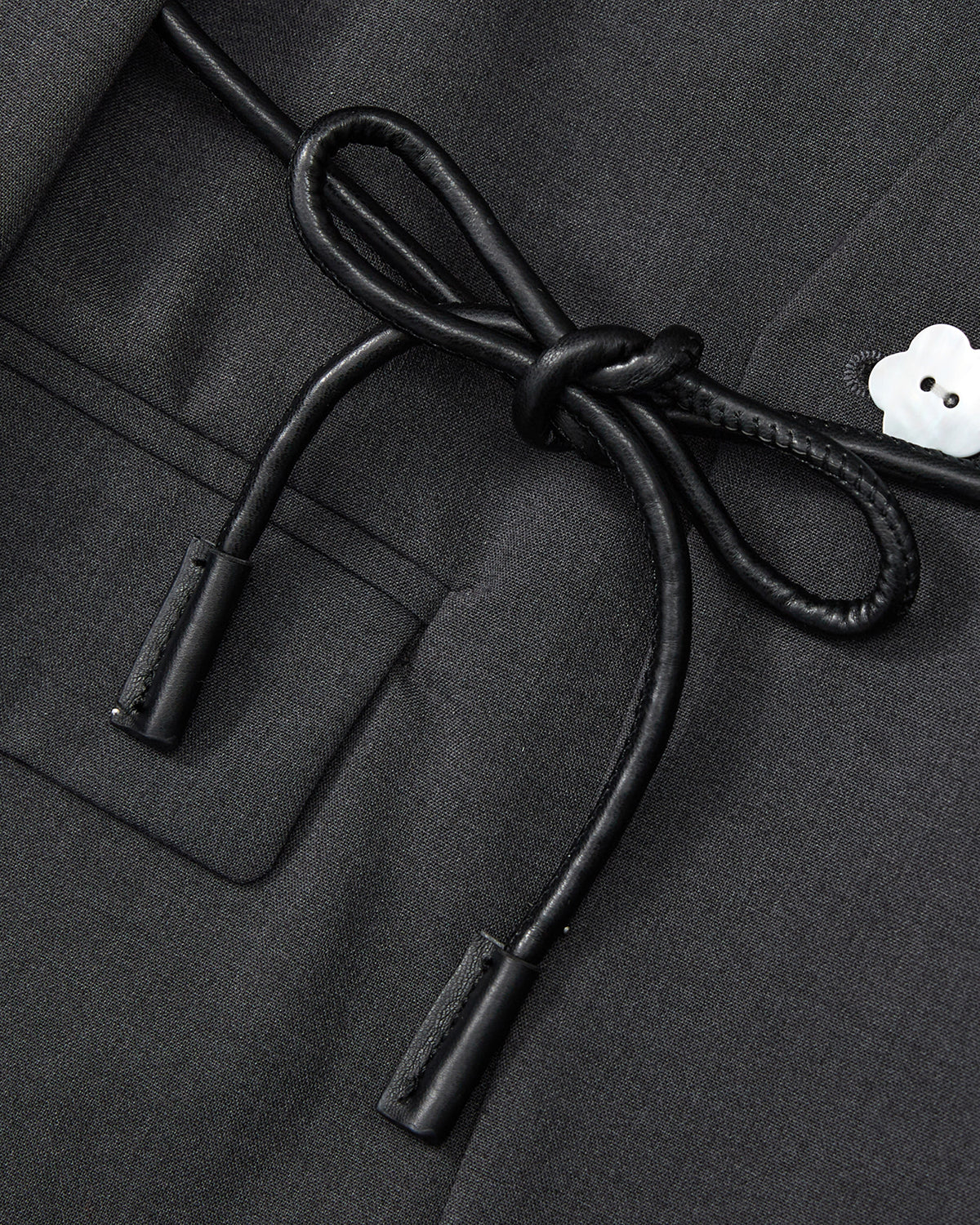 Close-up of the Sundae School COLLARLESS TIE BLAZER in dark gray, showing its breathable fabric, pocket detail, black braided tie bow, and a small white flower-shaped button.
