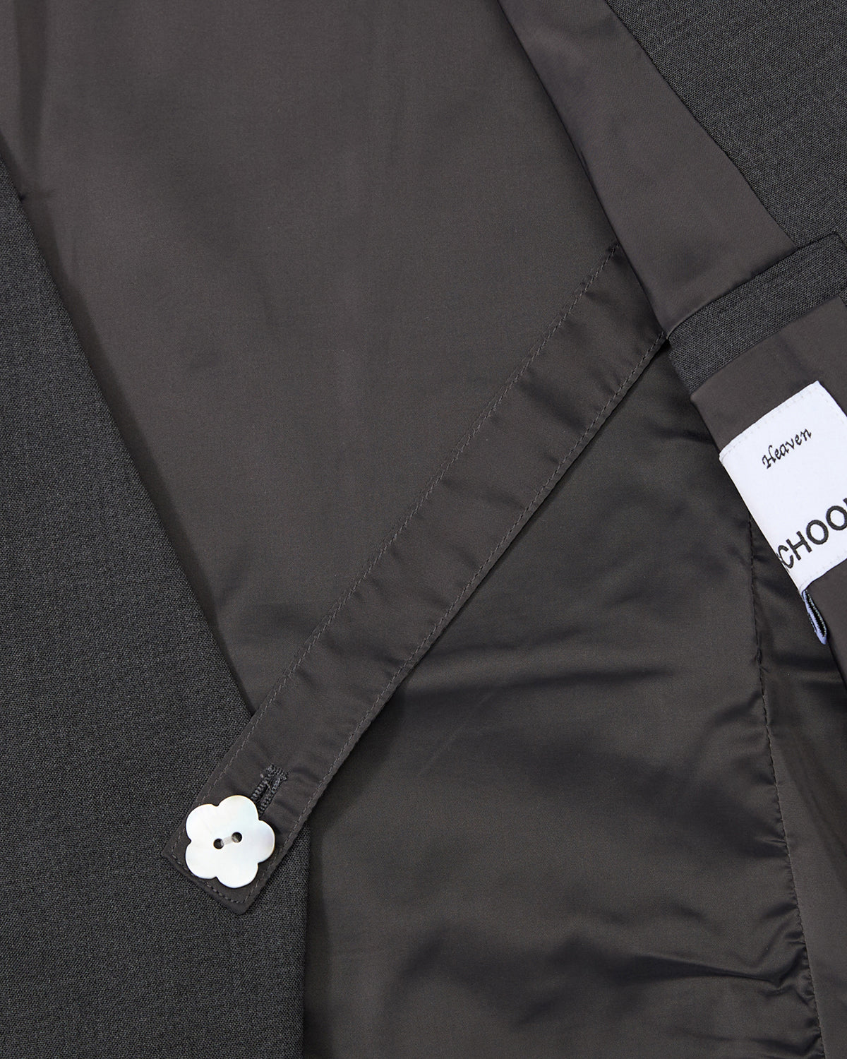 A close-up of the Sundae School COLLARLESS TIE BLAZER in dark grey highlights its inner lining, a fabric strap with a white flower button, and a partially visible label.