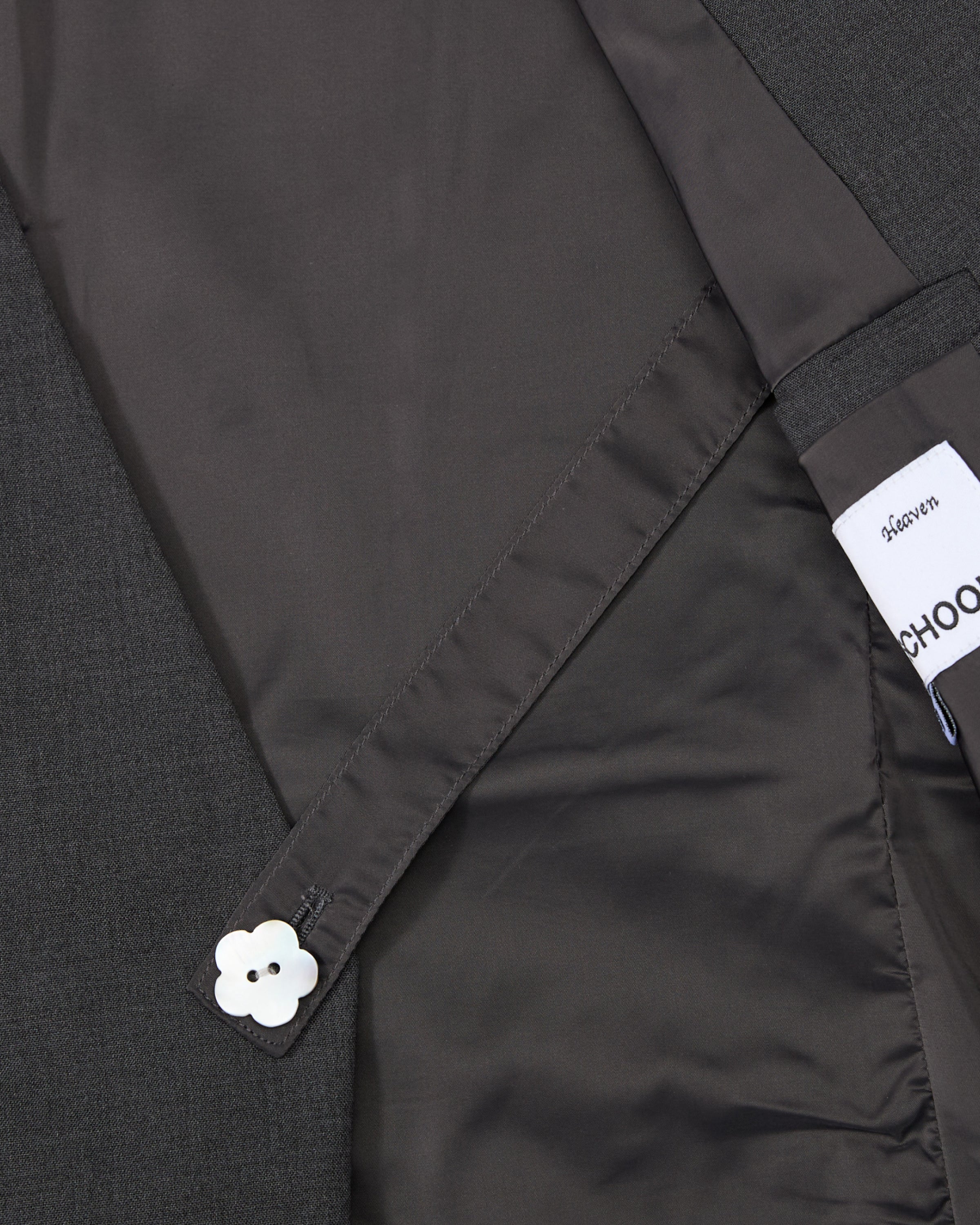 A close-up of the inside lining of Sundae School’s COLLARLESS TIE BLAZER in dark gray, featuring a buttoned strap with a white flower-shaped button and a partially visible Sundae School label, highlighting the blazer’s unstructured drape.