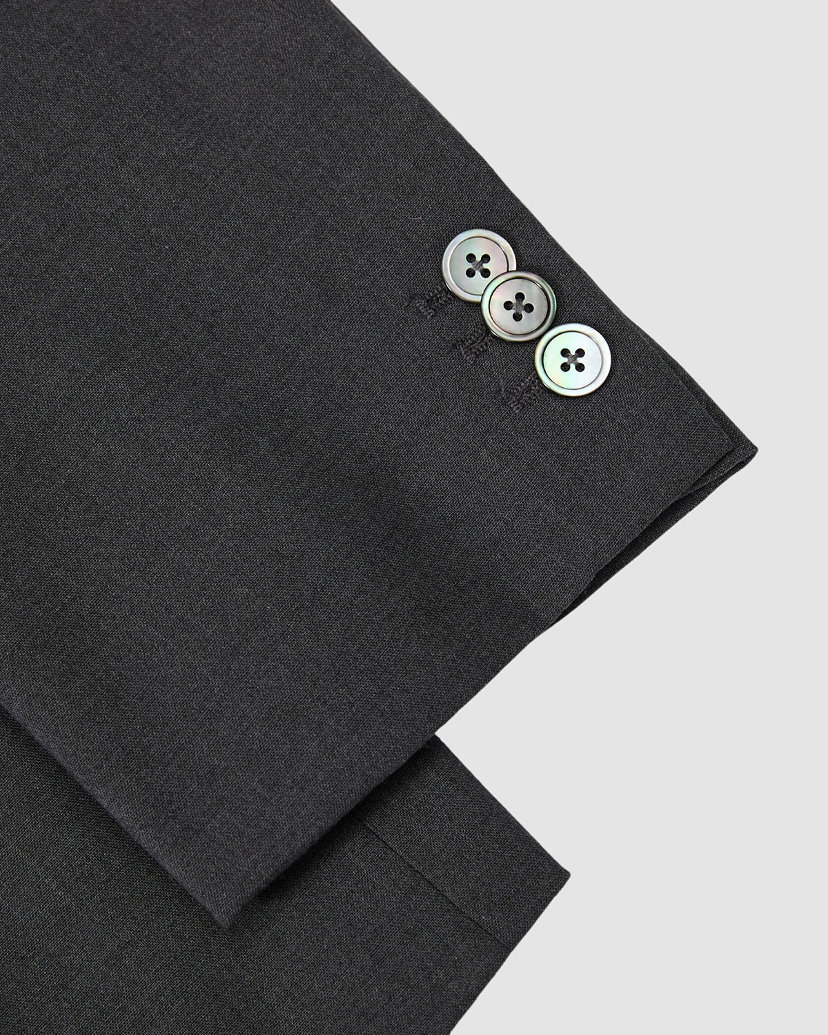 Close-up of the cuff on Sundae School’s COLLARLESS TIE BLAZER in dark gray, showing three shiny buttons on a breathable sleeve against a plain light gray background.